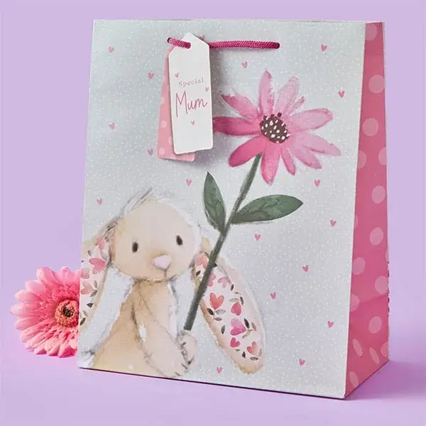 A pastel gift bag featuring a cute bunny holding a large pink flower, decorated with tiny hearts and a pink polka‑dot side panel, with a matching tag and a pink flower placed beside it.
